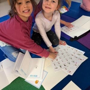 Two children placing yoga pose stickers into chakra-colored tracking books during a Grounded Kids Yoga class