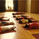 Twelve kids in Savasana (Resting Pose) lying quietly on yoga mats with eye pillows in a sunlit studio