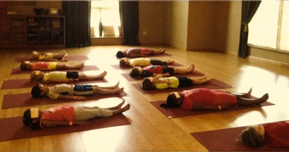 Twelve kids in Savasana (Resting Pose) lying quietly on yoga mats with eye pillows in a sunlit studio
