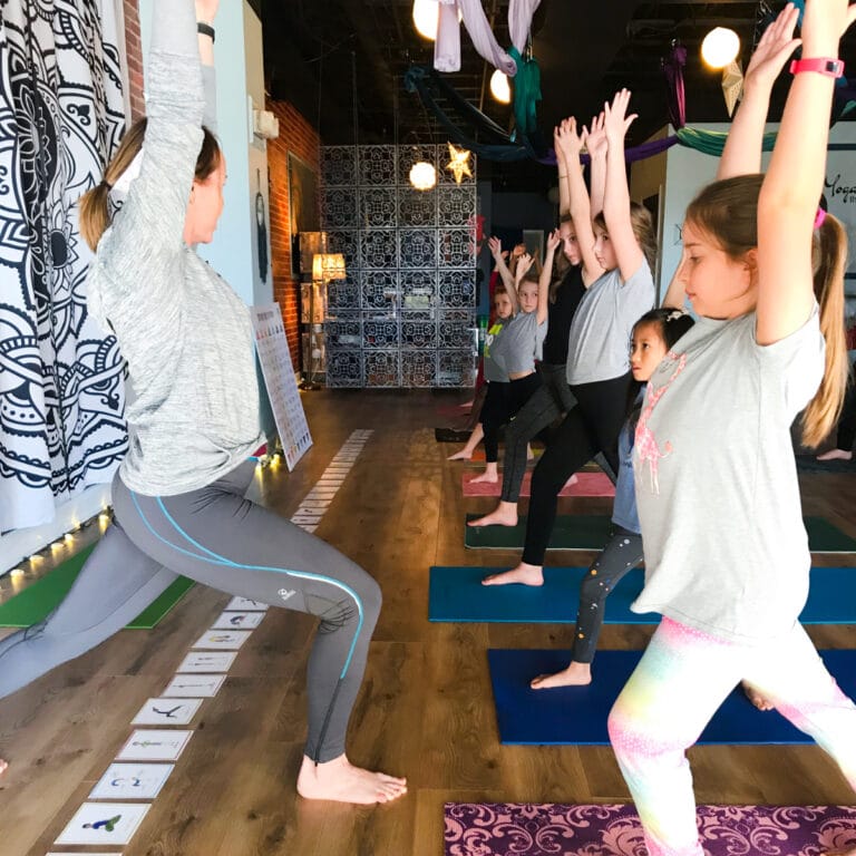 Group of children and adults practicing yoga together indoors with pose cards visible on the floor, part of Grounded Kids Yoga training