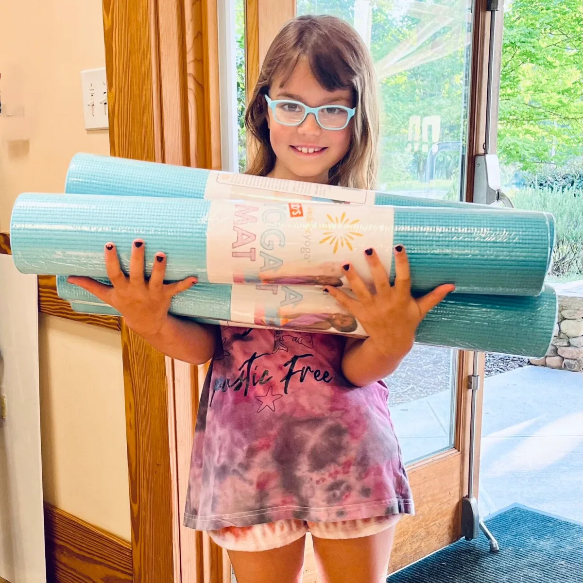 Child smiling and holding a rolled yoga mat, ready for an after-school Grounded Kids Yoga club session