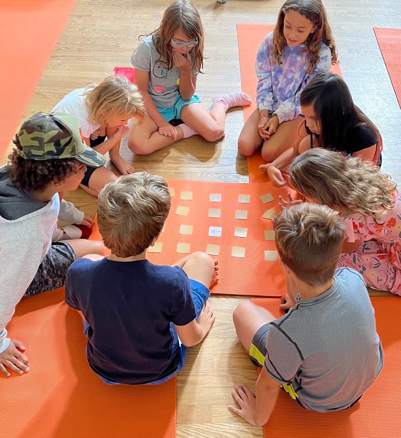 A group of homeschool children play a Grounded Kids Yoga memory game on orange mats, practicing focus and visual recall as part of a movement-based learning session