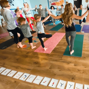 A group of trainees and children practicing yoga in a circle while holding hands during Step 4 of Grounded Kids Yoga certification, with pose cards on the floor