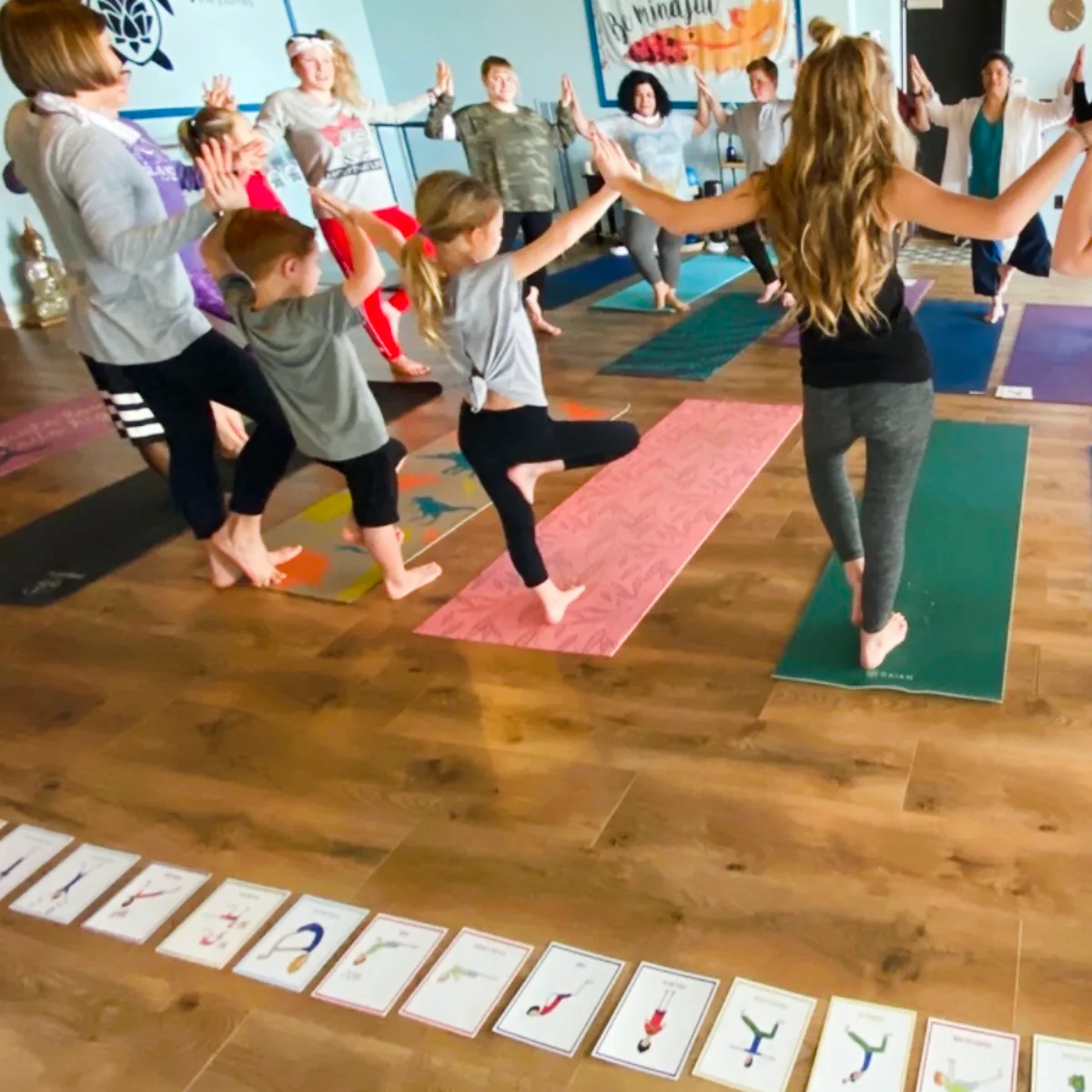 A group of trainees and children practicing yoga in a circle while holding hands during Step 4 of Grounded Kids Yoga certification, with pose cards on the floor