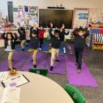 Children practicing Warrior I pose during a Grounded Kids Yoga class on purple mats in a school classroom