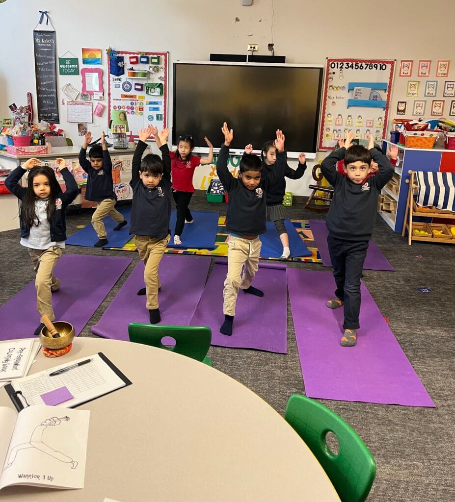 Children practicing Warrior I pose during a Grounded Kids Yoga class on purple mats in a school classroom