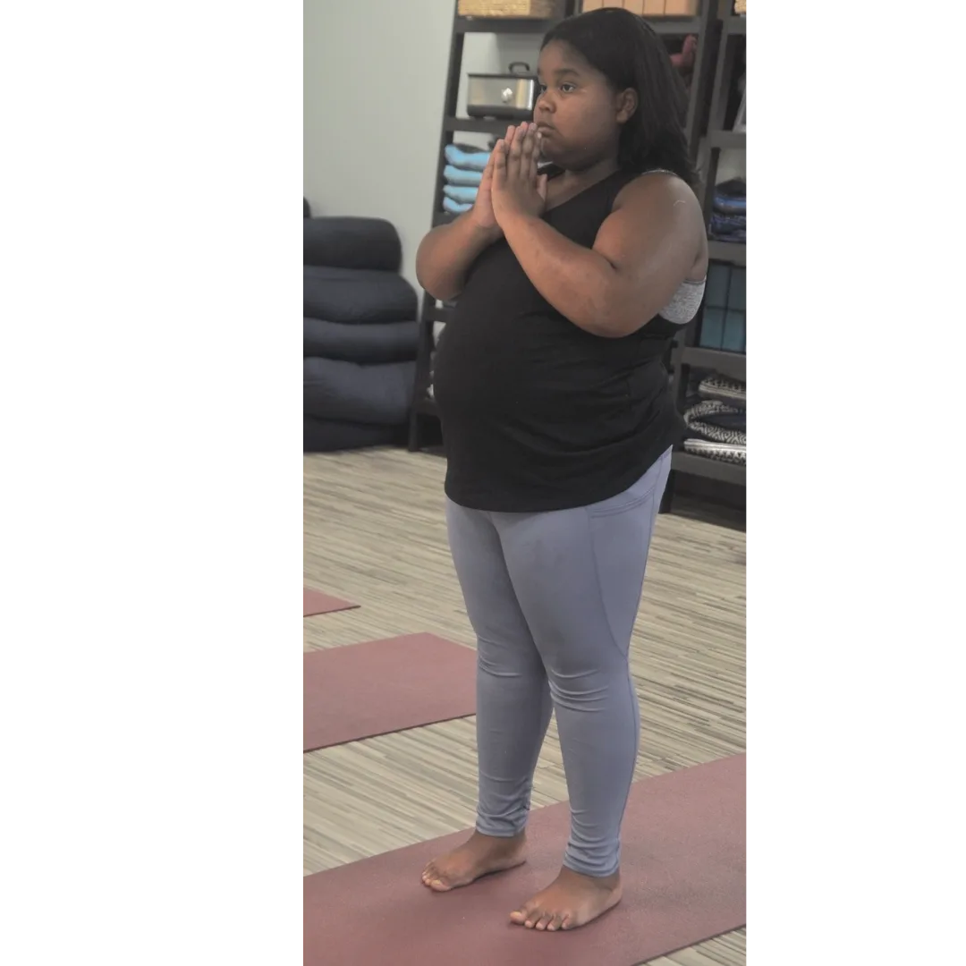 A young girl standing barefoot on a yoga mat in mountain pose with hands pressed together at her chest in a calm, focused posture.