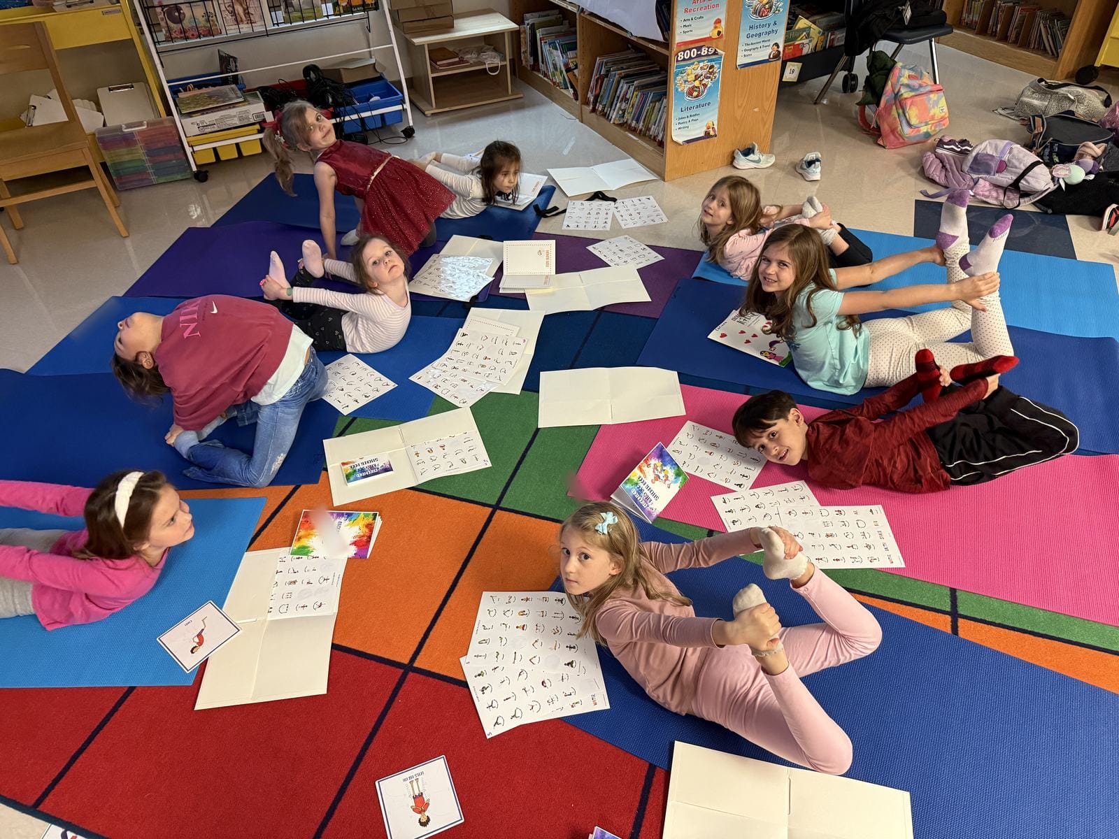 Children in a school library practising a backbend on yoga mats, with Grounded Kids pose charts, flow cards and sticker books spread on the floor around them