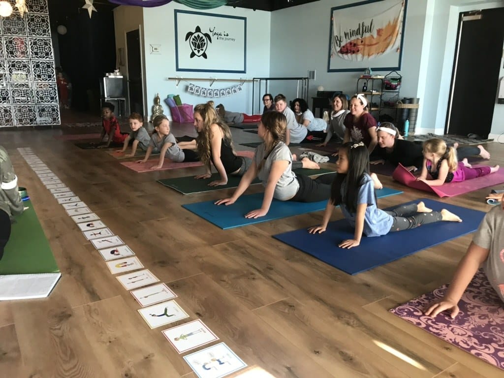 Children practising Cobra Pose in a kids yoga class at a host studio while adult trainees observe during a Grounded Kids teacher training.