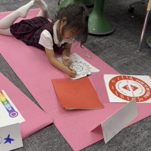 Girl in a plaid school uniform lying on a pink yoga mat coloring a mandala page, with the red Dare To Be Visible chakra color poster on the mat beside her