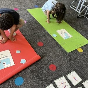Two children crouched on red and green yoga mats, each with a Grounded Kids Bingo board marked with blue paper squares. Green-bordered pose cards are spread on the carpet in front.