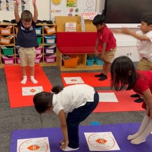 Children in school uniforms practicing yoga poses in a classroom, bending forward over red Grounded in Color Pose Clocks laid on purple and red yoga mats, as one boy stands with his arms raised.