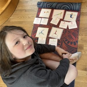Girl in a gray hoodie sitting on a patterned yoga mat at home beside a routine of Grounded Kids Yoga Pose Cards she laid out