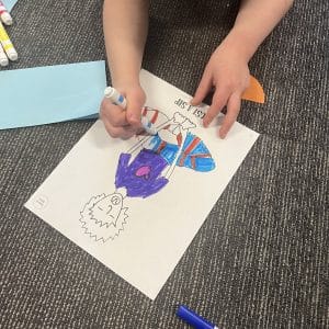 Close up of a child coloring the Just a Sip pose page from a Grounded Kids kids yoga coloring book with markers on a classroom carpet