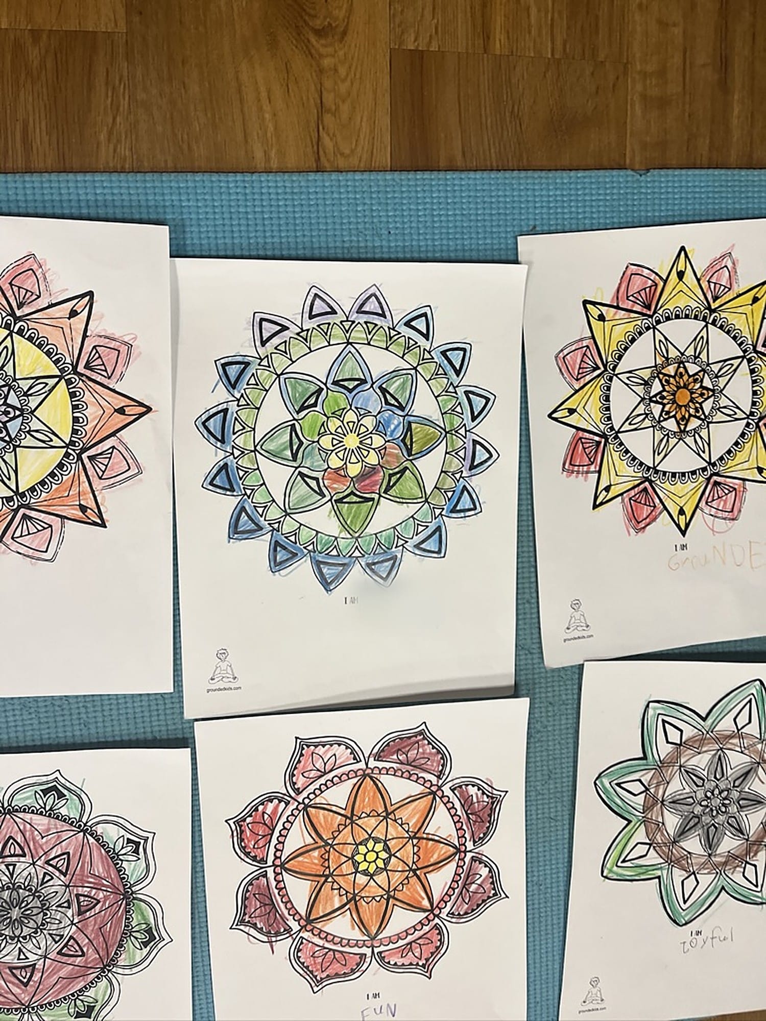 Six mandala pages from the I Am Centered Mandala Coloring Book, colored by kids in a Grounded Kids class, laid out on a teal yoga mat with "I Am" statements such as grounded, fun and joyful written under the designs.