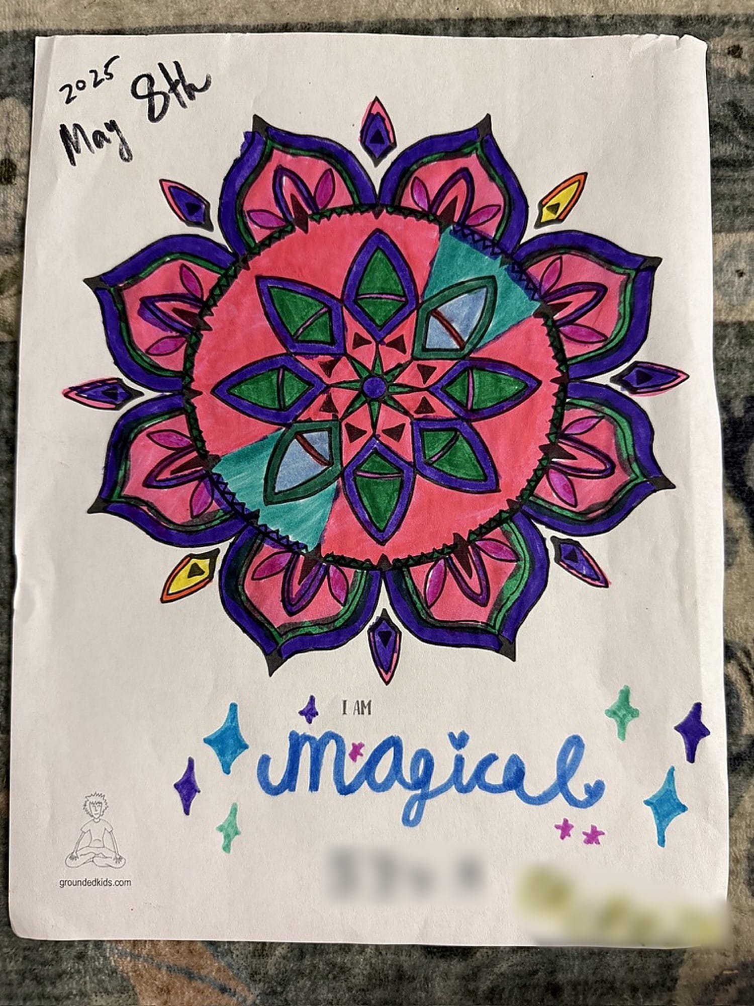 A mandala page from the I Am Centered Mandala Coloring Book colored in pink, purple and green by a child in a Grounded Kids class, with the "I Am" statement "magical" written under the design and surrounded by drawn stars.