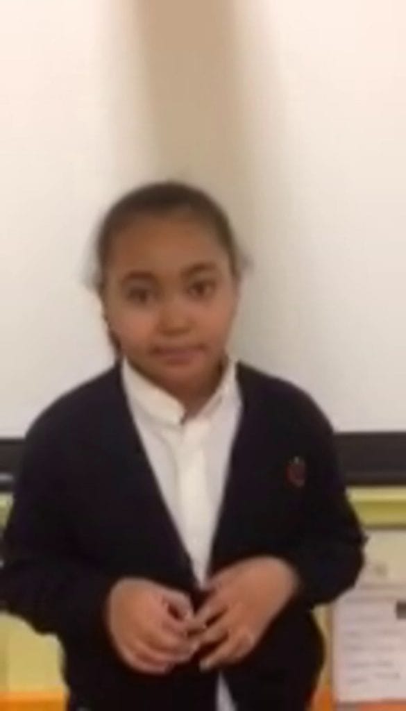 Fourth grade girl in a dark cardigan speaking to the camera, a still from the I Am What I Am video