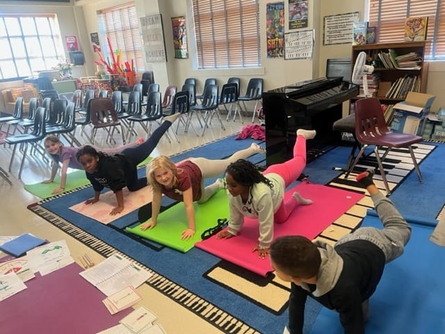 Children on hands and knees on yoga mats in a school music room, each with one leg stretched back, with pose cards and worksheets laid out on a mat in front.