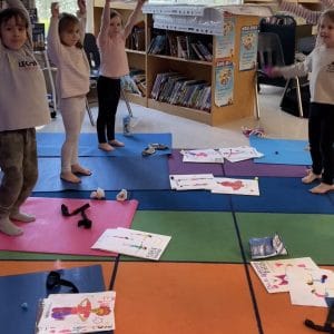 Children standing on yoga mats in a school library, most with their arms raised overhead, with pages from the Kids Yoga Coloring Book Bundle that they colored in laid on the mats and rug around them.