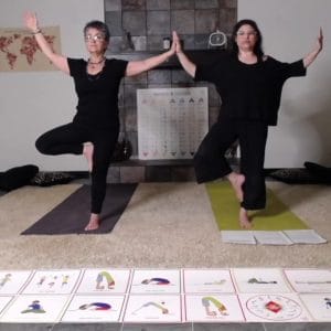 Amy and Sedef of Grounded Kids Yoga balancing in tree pose with palms joined in the Class 2 Grounding lesson video, pose cards laid out on the floor in front