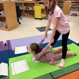Two children practicing a partner pose on yoga mats in a school library. One lies face down while the other stands over her holding her wrists, with the Kids Yoga Pose Poster on the mats beside them.