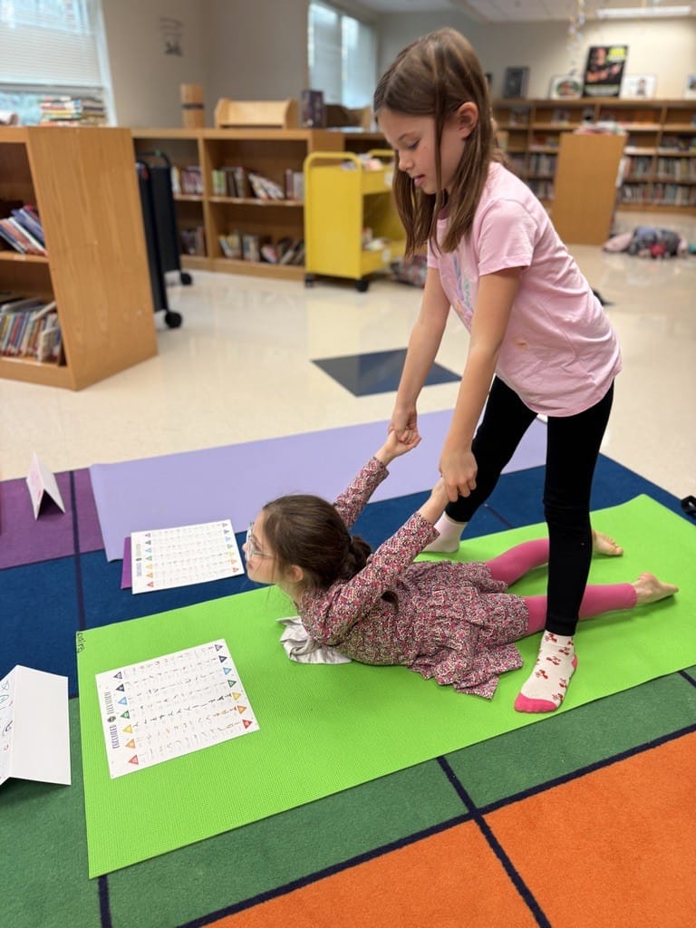 Two children practicing a partner pose on yoga mats in a school library. One lies face down while the other stands over her holding her wrists, with the Kids Yoga Pose Poster on the mats beside them.