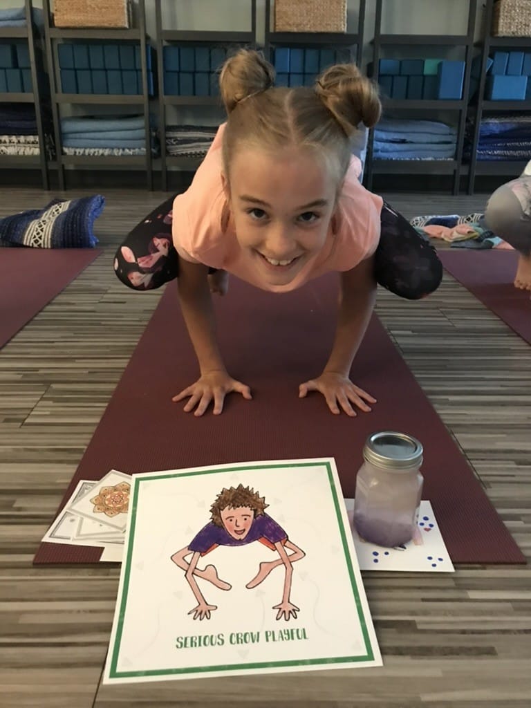 Smiling girl in crow pose on a maroon yoga mat behind the Serious Crow Playful card from the Grounded Kids Yoga Pose Cards deck