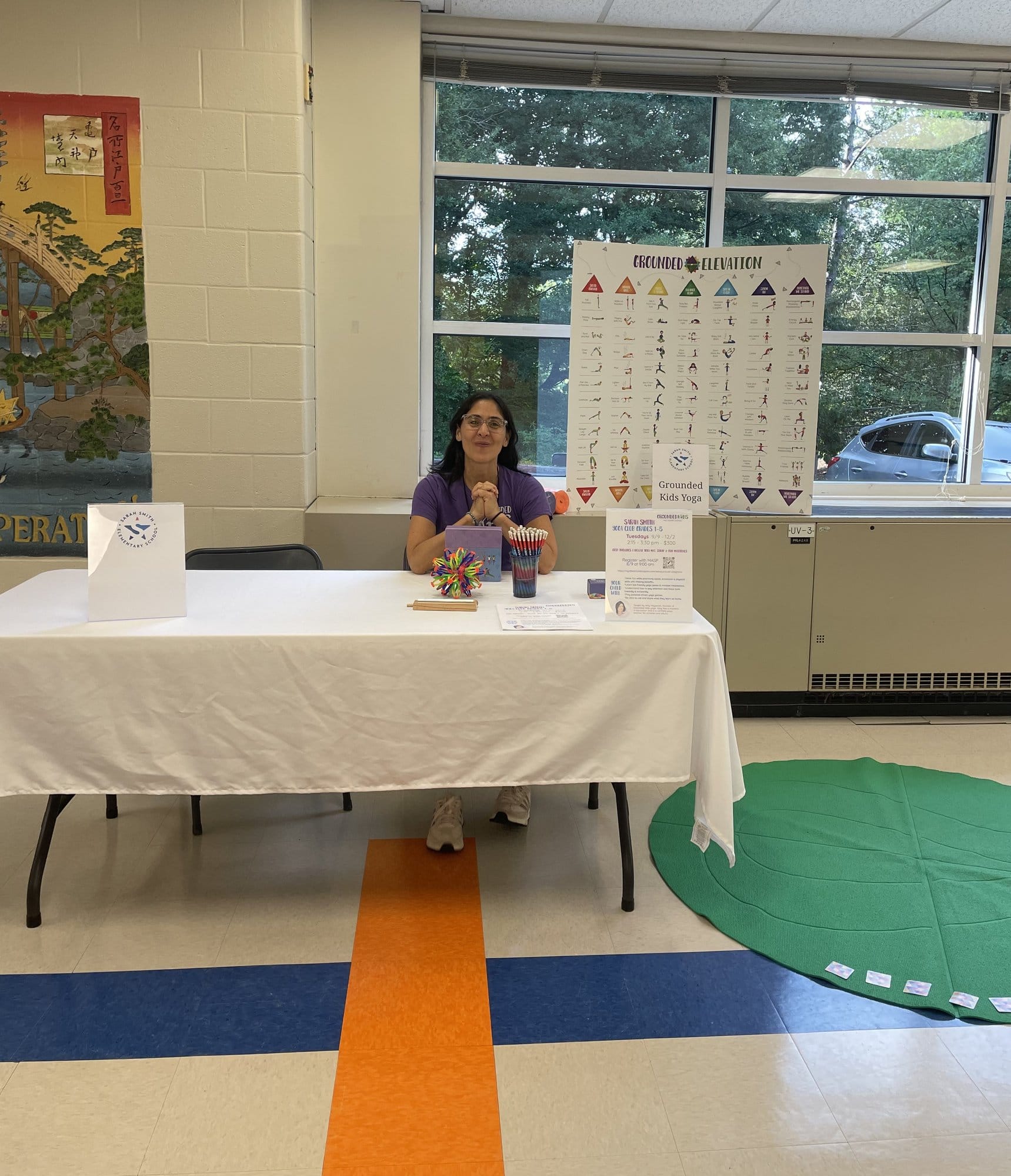 A woman seated at a Grounded Kids Yoga table covered in a white cloth, with the Grounded Elevation pose poster on a stand behind her.
