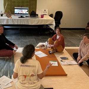 Grounded Kids founder Amy teaching Step 1 in person, seated with trainees on yoga mats, with online trainees on the Zoom screen behind.