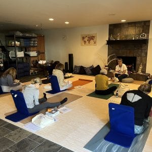 Grounded Kids lead trainer Sedef teaching Step 3 from the front of the studio by the fireplace, with trainees seated on yoga mats and floor chairs taking notes.