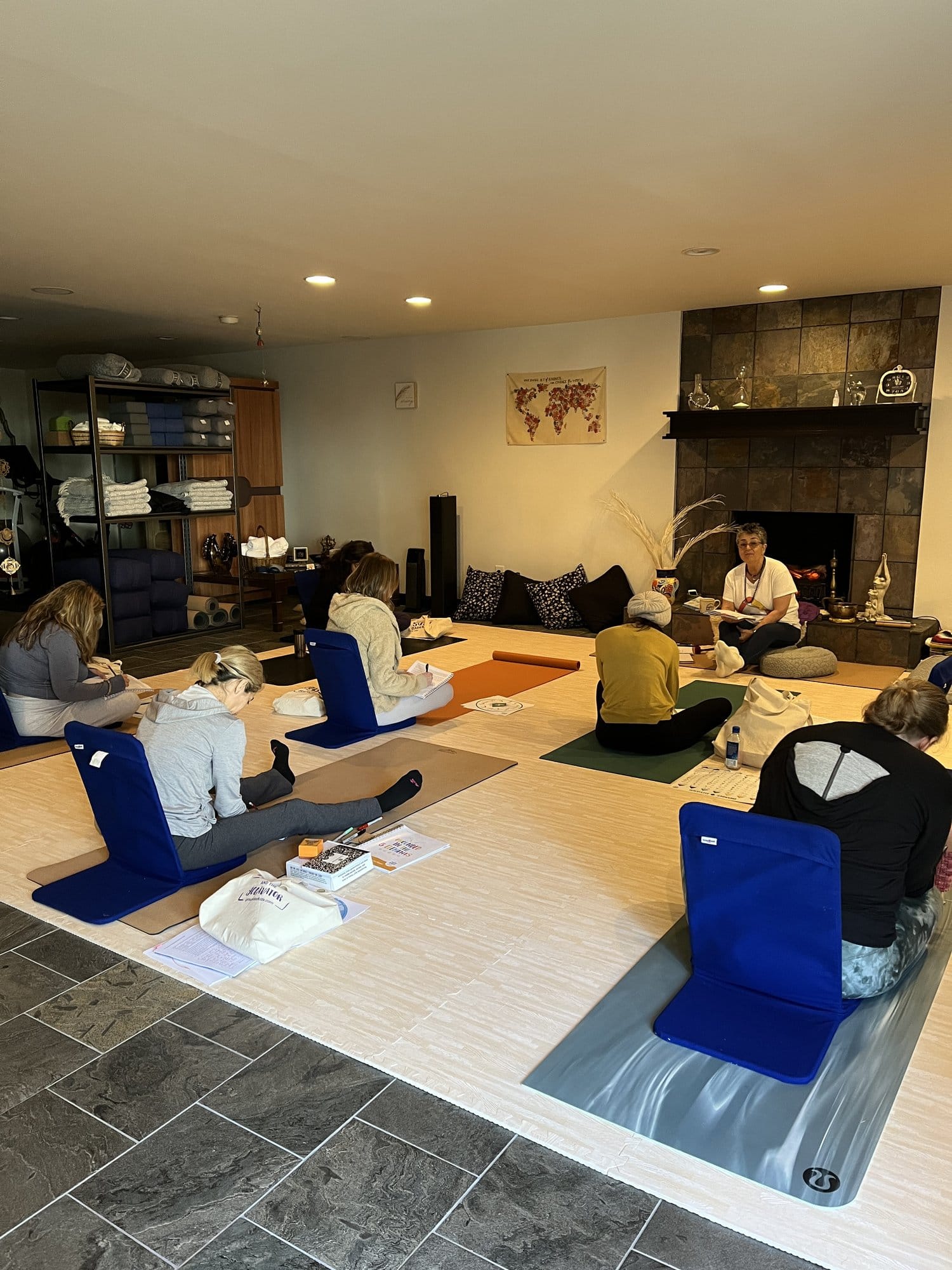 Grounded Kids lead trainer Sedef teaching Step 3 from the front of the studio by the fireplace, with trainees seated on yoga mats and floor chairs taking notes.