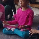 Girl in a pink shirt sitting cross-legged with her eyes closed and her hands resting on her knees during a kids yoga class