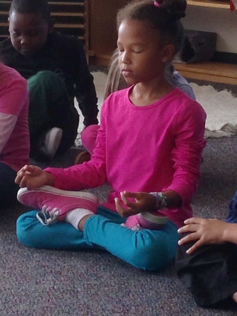 Girl in a pink shirt sitting cross-legged with her eyes closed and her hands resting on her knees during a kids yoga class