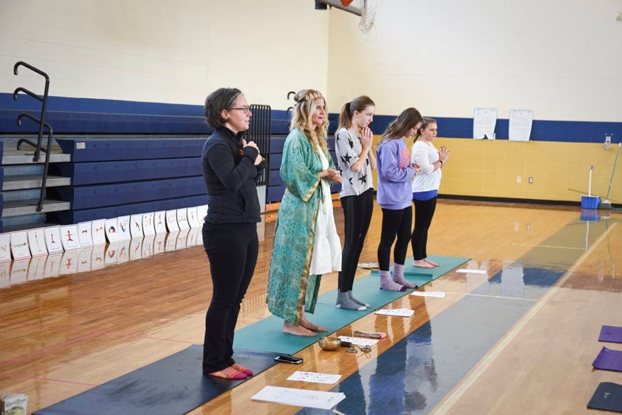 Teacher in a green robe and adults standing with hands at the heart in a school gym