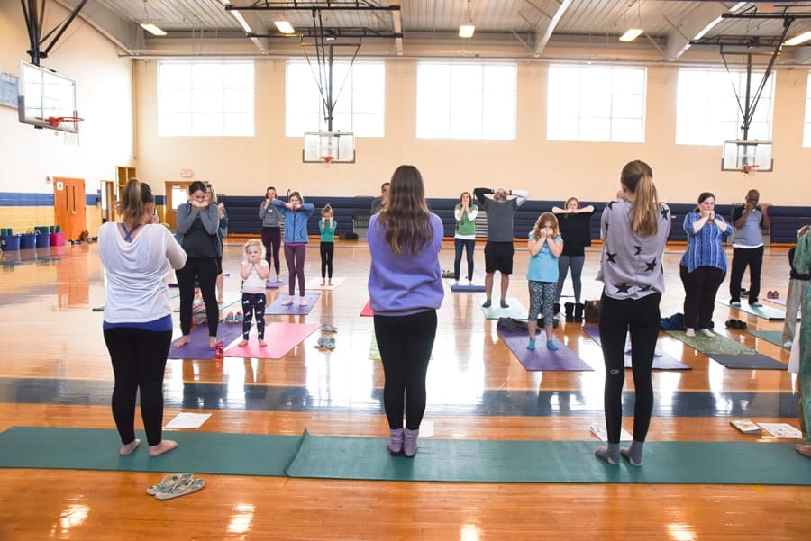 Adults and children standing on yoga mats facing the front of a school gym