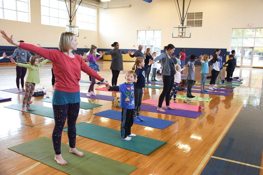 Adults and children standing on yoga mats with arms stretched out to the sides in a school gym