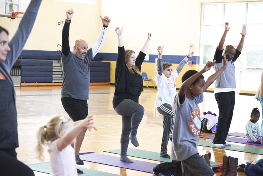 Adults and children balancing on one leg with arms raised overhead in a school gym