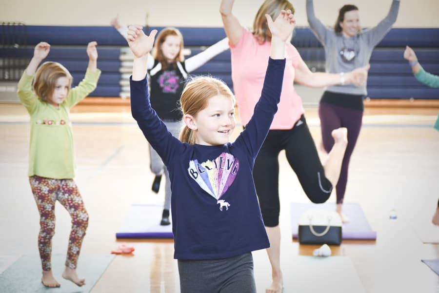 Smiling girl balancing on one leg with arms raised overhead beside adults