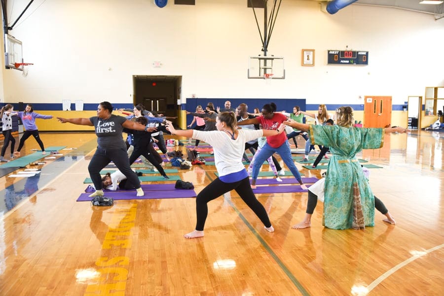Adults and children in Warrior II across a school gym, a teacher in a green robe among them