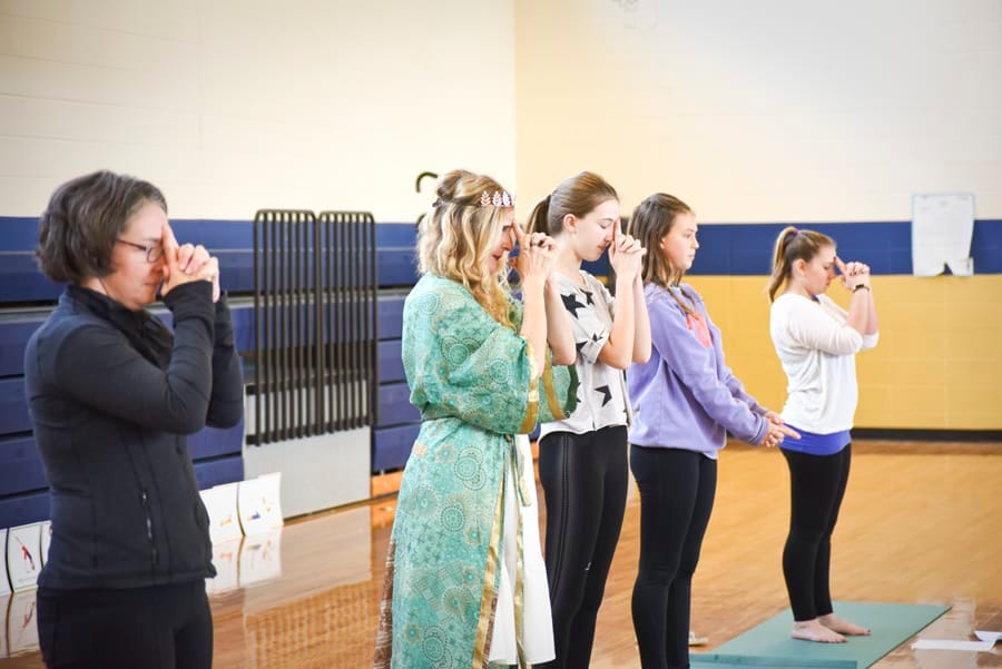 Teacher in a green robe and girls standing in a row with hands raised to their faces