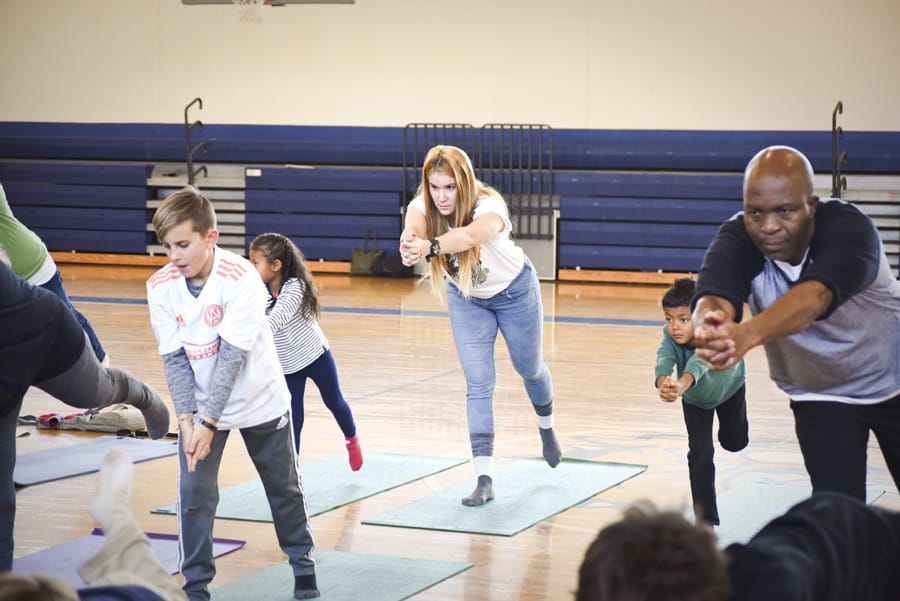 Adults and children balancing on one leg and leaning forward in a school gym