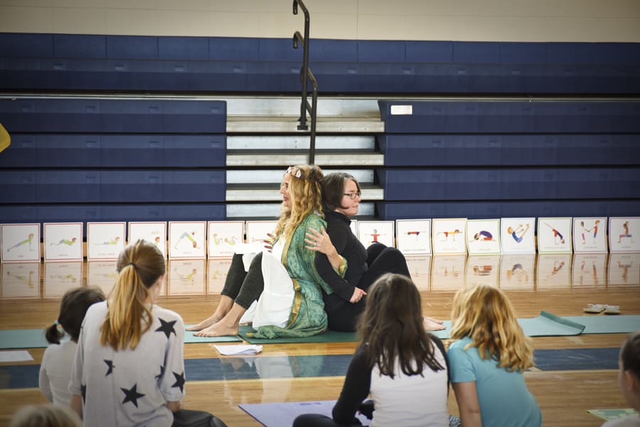 Teacher in a green robe seated at the front of a school gym with children sitting facing her
