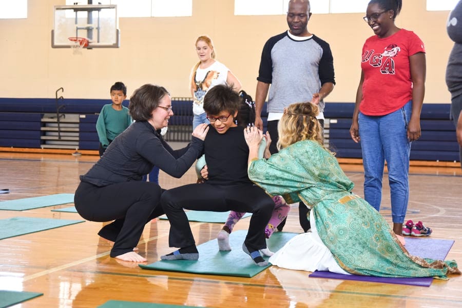 Teacher in a green robe and a woman crouching beside a child on a mat while adults watch