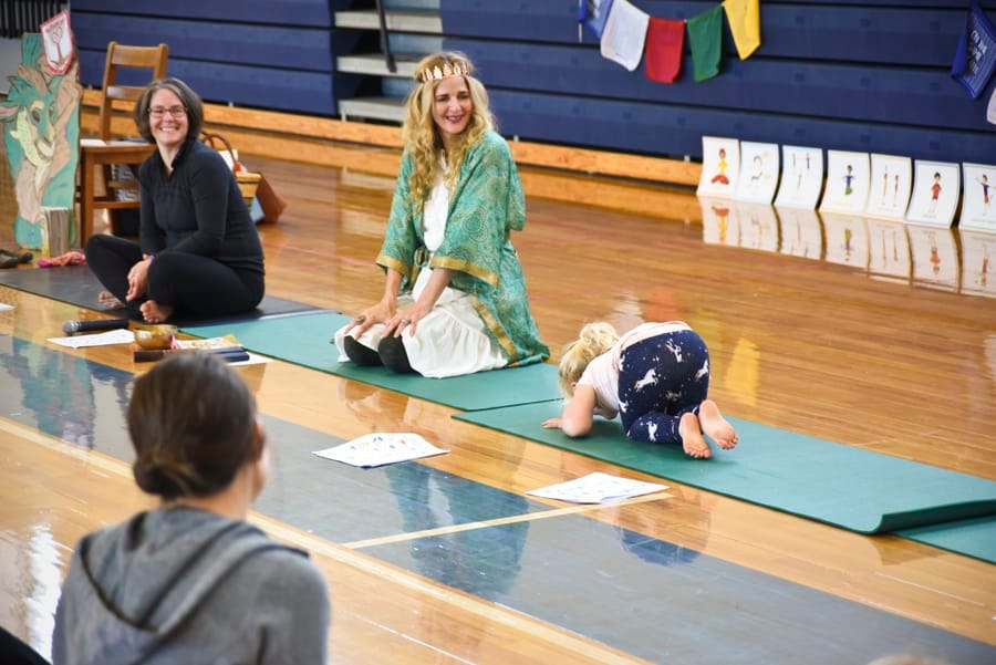 Teacher in a green robe kneeling at the front of a school gym as a child rests in child's pose
