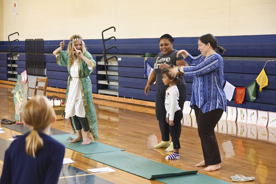Teacher in a green robe leading women and a child standing on one leg in a school gym