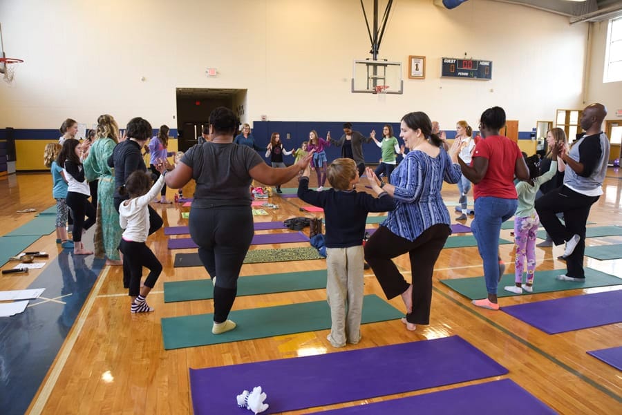 Adults and children holding hands in a circle, balancing on one leg in a school gym
