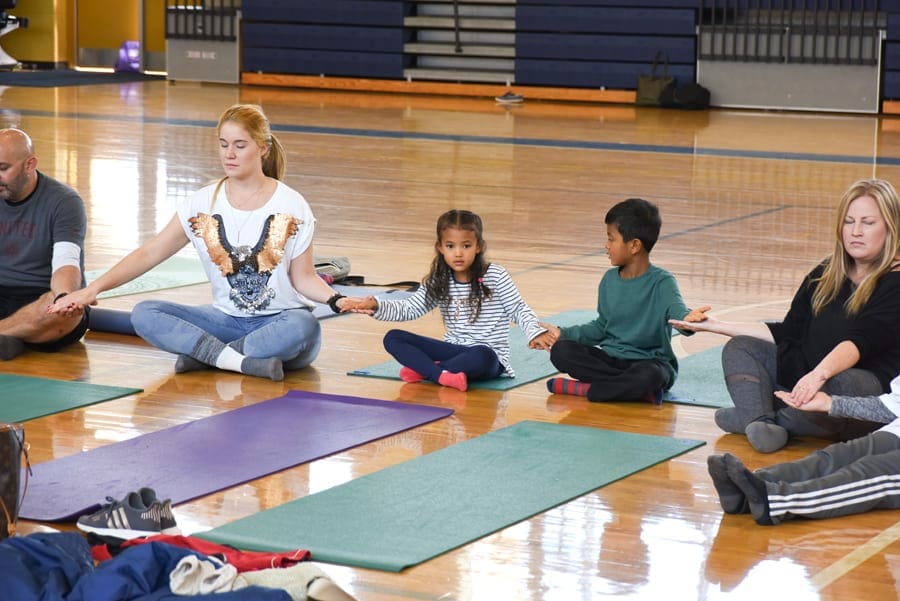 Adults and children sitting cross-legged in a circle, holding hands on yoga mats