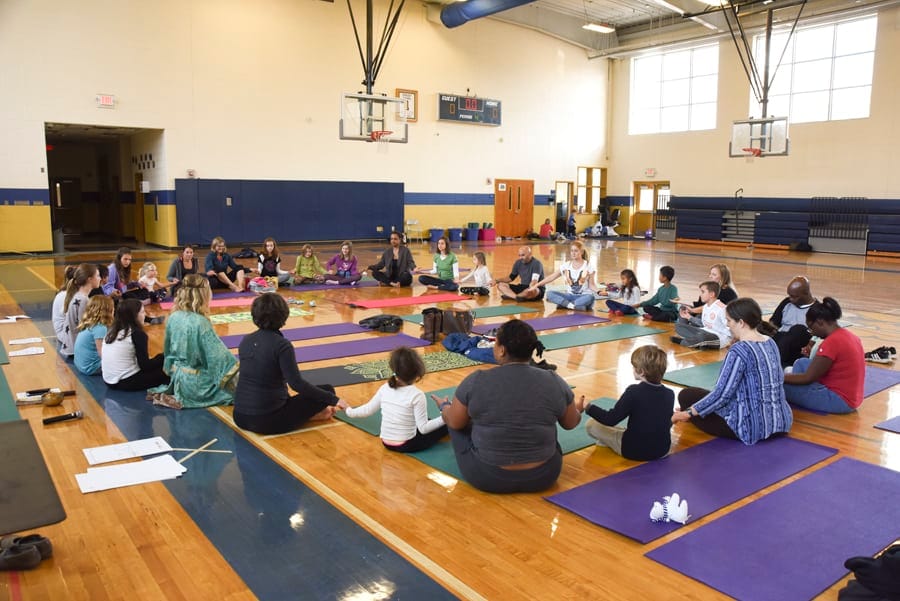 Large group of adults and children sitting in a circle on yoga mats in a school gym
