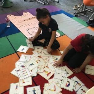 Two children sitting on a colorful classroom rug sorting Grounded Kids Yoga Pose Cards spread across the floor