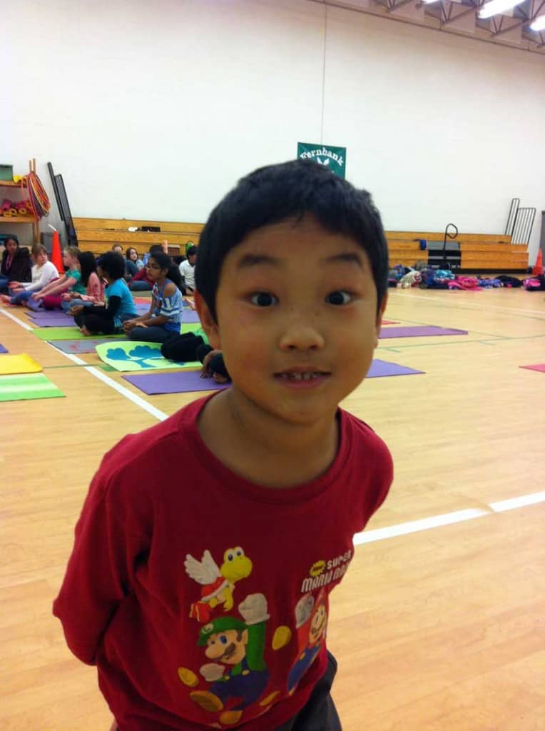 Smiling boy in a red t-shirt in a school gym, with children on yoga mats behind